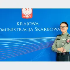 Kobieta w mundurze służby celno-skarbowej stoi przed niebieskim tłem z napisem "Krajowa Administracja Skarbowa".