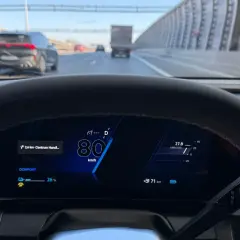 Widok z kabiny samochodu na autostradzie. Na wyświetlaczu 80 km/h, co jest minimalną prędkością na autostradzie. Widać inne pojazdy i barierki.