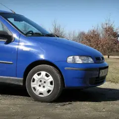 Błękitny Fiat Albea stoi na polnej drodze. W tle widać drzewa i błękitne niebo.