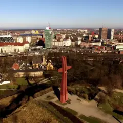 Wielki czerwony krzyż na górze gradowej góruje nad miastem. W tle widać zabudowania i drapacze chmur.