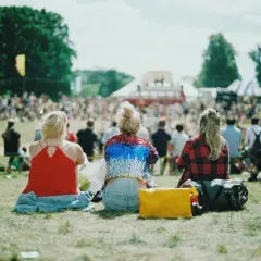 Festivalgäster sitter på gräset med en packlista i åtanke. En gul väska och en stor bag ligger redo för äventyr.