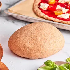 Ein runder Laib vollkorn pizzateig liegt bereit. Im Hintergrund eine fast fertige Pizza mit Tomaten und Mozzarella.