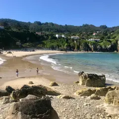 La **playa de Amió** es un paraíso de arena dorada y aguas cristalinas, con colinas verdes y casas blancas al fondo.
