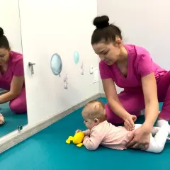 Fizjoterapeutka wspiera niemowlę na brzuchu, stosując metodę PNF. Dziecko ćwiczy podnoszenie główki, a terapeuta delikatnie koryguje jego ułożenie.