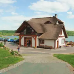 Dom z piękną, naturalną strzechą na dachu, położony nad jeziorem. Wokół zielony trawnik, drewniany wóz i rzeźby.