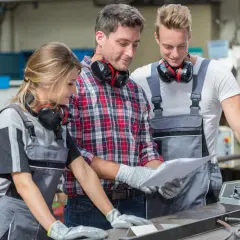 Drei SHK-Techniker besprechen Pläne. Sie tragen Arbeitskleidung und Kopfhörer. Das Gehalt eines SHK-Technikers ist ein wichtiges Thema.