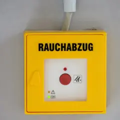 Gelbes Gehäuse für Rauchabzug mit rotem Knopf. Ein grünes Licht zeigt "OK" an, was auf ein erfolgreiches Wartungsprotokoll RWA Muster hindeutet.