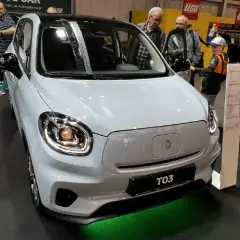 Biały, miejski samochód elektryczny MG T03 na wystawie. W tle widać innych zwiedzających i stoisko LEGO.