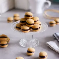 Baci di dama, deliziosi biscotti con crema al cioccolato, disposti su un'alzata di vetro, circondati da libri e tazze.