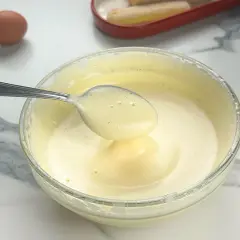 Préparation d'un tiramisu : une cuillère de pâte à bombe aérienne est prélevée d'un bol en verre. Un œuf et des biscuits à la cuillère sont visibles.