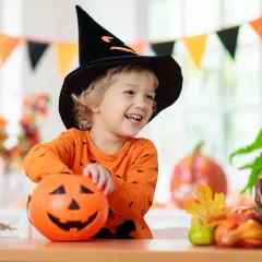 Uśmiechnięty chłopiec w czarnej, spiczastej czapce czarownicy i pomarańczowej bluzce z nietoperzami trzyma dynię. To idealny strój owoc dla dziecka na Halloween!