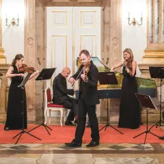 Ensemblen ger en klassisk musik konsert i en elegant sal. Sex musiker spelar fioler, cello och cembalo.