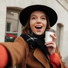 Uśmiechnięta dziewczyna w kapeluszu i płaszczu robi selfie z kawą. Chcesz wiedzieć, jak ustawić makijaż w aparacie iPhone?