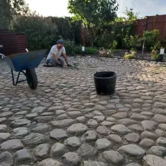 Mężczyzna pracuje przy podjeździe z kamienia polnego, obok stoi taczka i wiadro.