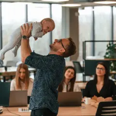 Mężczyzna w okularach podnosi dziecko do góry w biurze, w tle pracują kobiety.