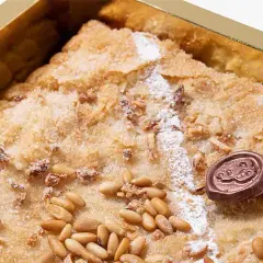 Deliciosa coca de Llavaneres con piñones y un toque de azúcar glas, presentada en una caja dorada.