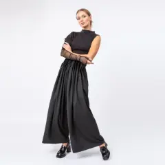 Kobieta w czarnym, eleganckim stroju z szerokimi spodniami i siateczkowymi rękawami, pozuje na białym tle.