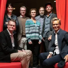 Gruppbild med åtta personer framför röda draperier. Izabella Jensen står i mitten, klädd i en mönstrad tröja.