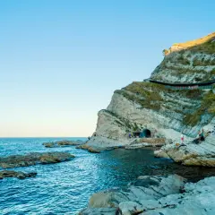 Scogliere rocciose e mare blu intenso al Conero, con un tunnel scavato nella roccia e persone che si godono la giornata.