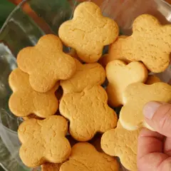 Una mano prende uno dei tanti biscotti fatti in casa a forma di quadrifoglio, disposti in una ciotola di vetro con foglie verdi sullo sfondo.
