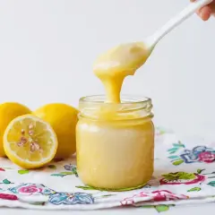 Cucharada de cremoso lemon curd cayendo en un frasco, junto a limones frescos sobre un paño floral.