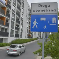 Znak "Droga wewnętrzna" informuje o strefie zamieszkania. Widać tu pieszych, samochód i dom.