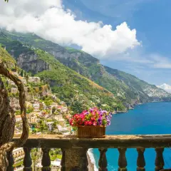 A vibrant view of Amalfi Coast towns clinging to steep green hillsides, overlooking the sparkling blue sea. A twisted tree frames the scene, with a planter of colorful flowers on the balcony railing.