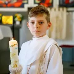 Chłopiec w białej komży trzyma ozdobną świecę podczas I Komunii Świętej.