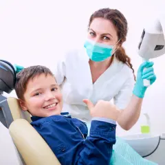 Uśmiechnięty chłopiec daje kciuk w górę w gabinecie stomatologicznym. Uśmiechnięta dentystka w masce i rękawiczkach stoi za nim.