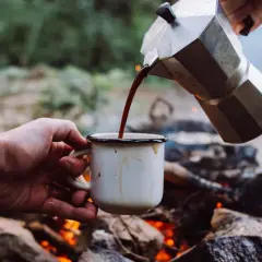Gorąca kawa w górach, nalewana z kawiarki do kubka nad ogniskiem.