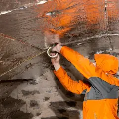 Travaux d'isolation dans une cave en pierre. Un ouvrier en combinaison orange fixe un isolant réfléchissant au plafond avec du ruban adhésif.