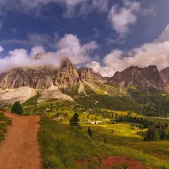 Camino de tierra rojiza que se adentra en un valle alpino con imponentes montañas cubiertas de nubes. La ruta Cinque Torri se revela entre prados verdes y bosques frondosos.