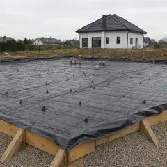 Przygotowane wykopy pod fundamenty, zbrojenie i izolacja przeciwwilgociowa czekają na beton. W tle domy jednorodzinne.