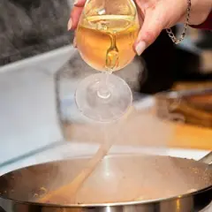 Eine Hand gießt Weißwein aus einem Glas in eine dampfende Pfanne. Das Verkaufen von Wein beim Kochen verleiht dem Gericht Tiefe.