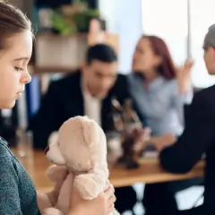 Smutna dziewczynka z misiem, podczas wizyty kuratora sądowego w domu. W tle rodzice i prawnik.