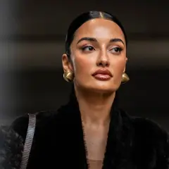 Mujer con peinado pulido y maquillaje natural, luciendo aretes dorados y un abrigo negro de pelo. Su **clean look** irradia elegancia.