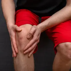Mężczyzna trzyma się za bolące kolano w czerwonych spodenkach. Zastanawia się, jak długo boli przeciążone kolano po treningu.