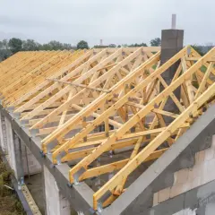 Drewniana konstrukcja dachu bez podbitki, widoczna na budowie domu. Widać komin i elementy ścian z bloczków.