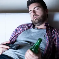 Mężczyzna śpi na kanapie z pilotem i butelką piwa, być może doświadczając dziwnych snów po odstawieniu alkoholu.