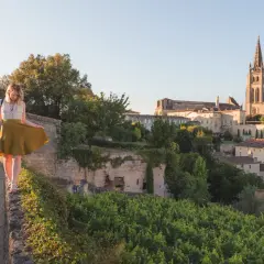 Une femme en jupe jaune marche sur un muret, surplombant le village de Saint-Émilion. La route des vins s'étend à perte de vue.