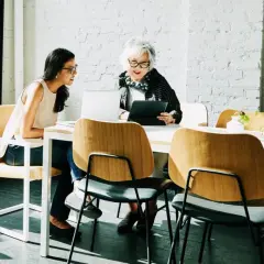 Zwei Frauen besprechen am Tisch die betriebsrente. Nachteile werden diskutiert, während sie auf Laptops und Tablets schauen.