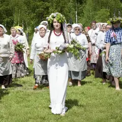 Grupa kobiet w tradycyjnych strojach i wiankach idzie przez łąkę. Centralna postać w białej sukni niesie bukiet kwiatów. Obraz nawiązuje do tradycji Zielonych Świątek.