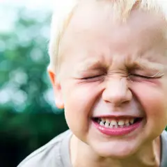 Zmarszczony chłopiec z blond włosami, zęby wyszczerzone w grymasie. Może to być reakcja na pytanie: dlaczego dziecko gryzie mamę?