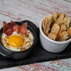 Un œuf cocotte raclette, crémeux et gourmand, garni de noix et de fines tranches de viande séchée, servi avec des toasts croustillants.