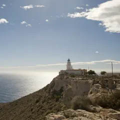 El faro de Mesa Roldán se alza sobre un acantilado rocoso, vigilando el mar Mediterráneo bajo un cielo azul con nubes dispersas.