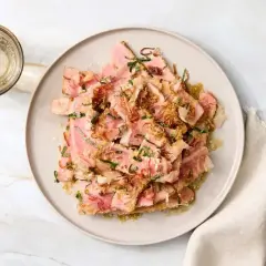 Tataki z ryby na talerzu, posypane ziołami i chrupiącą cebulką, obok szklanka napoju.