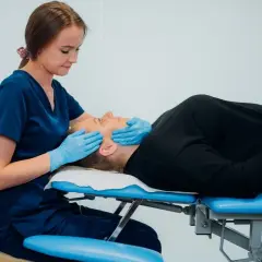 Fizjoterapeuta szczękowy delikatnie masuje twarz pacjentki leżącej na kozetce.