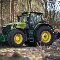 Traktor John Deere z charakterystyczną kratką w żółto-zielone wzory, wyposażony w mulczer do traktorka, pracuje w lesie.