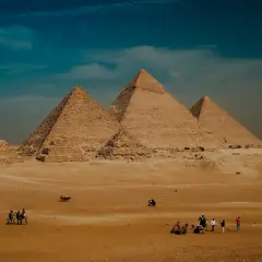 Pustynia Giza z piramidą Cheopsa w tle. Ludzie i wielbłądy przemierzają piaski pod błękitnym niebem.