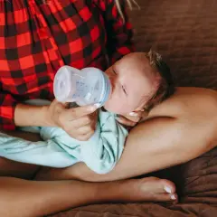 Mamma con maglia a quadri rossi offre il biberon a un neonato, praticando l'allattamento misto.